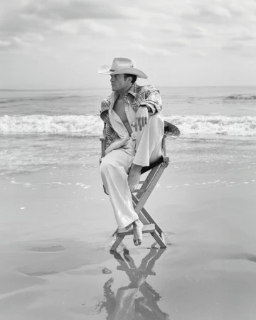 Orlando Bloom barefoot in a cowboy hat sitting on a director chair in shallow ocean surf on a sandy beach in black and white.