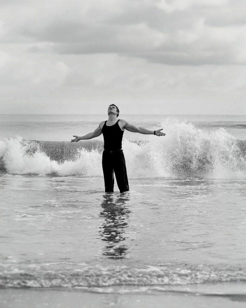 Monochrome photograph of a man in dark clothing standing in ocean water with arms outstretched under an overcast sky and crashing waves.