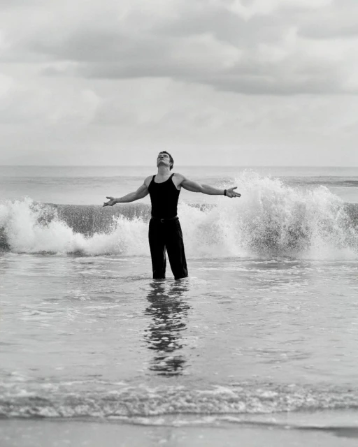 Monochrome photograph of a man in dark clothing standing in ocean water with arms outstretched under an overcast sky and crashing waves.