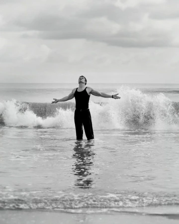 Monochrome photograph of a man in dark clothing standing in ocean water with arms outstretched under an overcast sky and crashing waves.