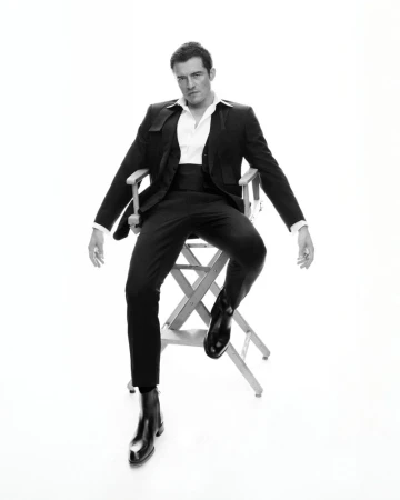 Orlando Bloom seated on a director's chair wearing a tailored black suit and white collared shirt against a white seamless backdrop.