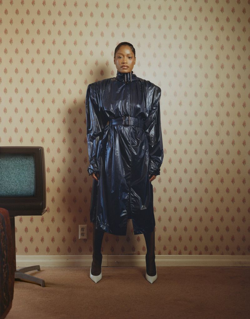 Keke Palmer wearing a glossy dark blue belted coat and white pumps standing against patterned wallpaper