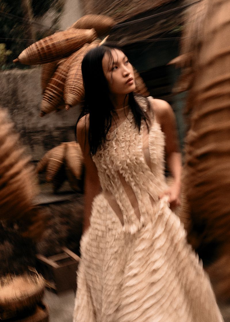 Woman in textured dress poses in front of woven objects and wall, medium shot, blurred motion, fashion, beauty, and editorial themes.