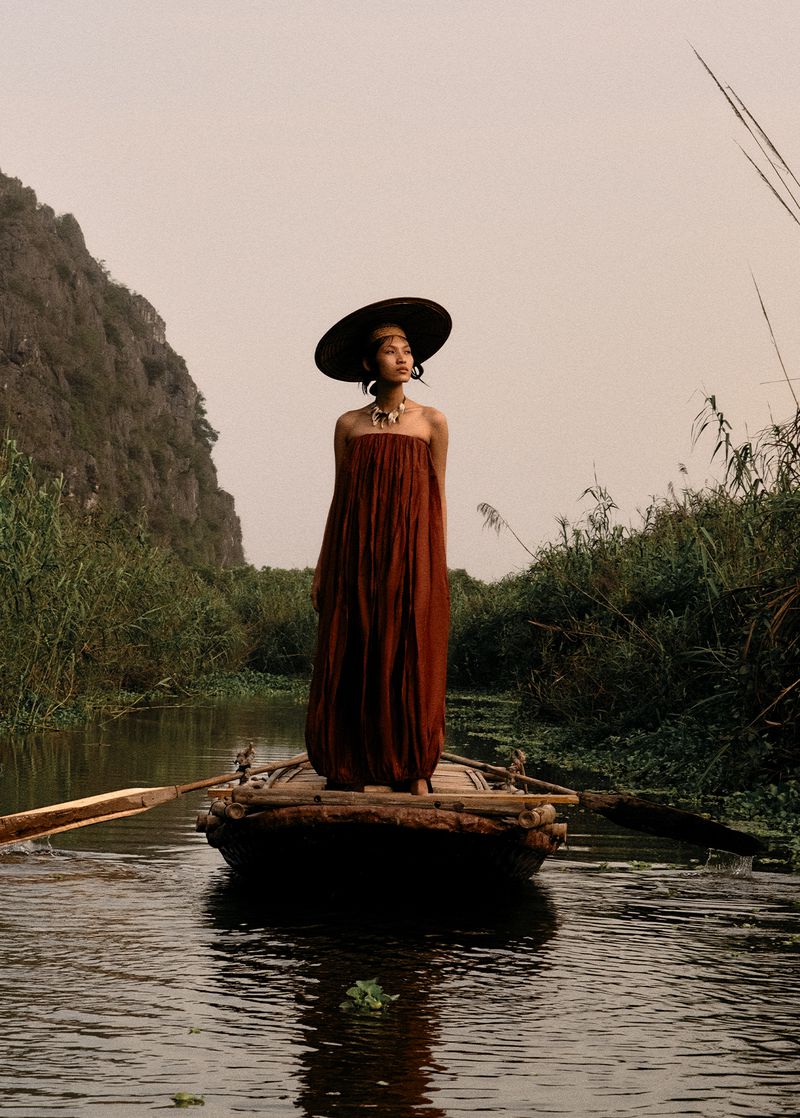 Woman wearing brown dress and large hat stands on a boat floating on a river surrounded by reeds and a mountain background.