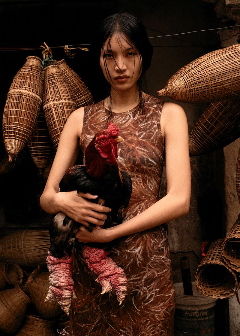 Portrait of Asian woman in brown dress holding a large rooster with legs and feet visible, surrounded by woven wicker baskets.