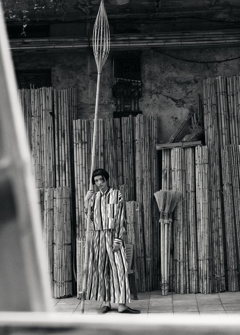 Monochrome portrait of an adult female in a striped suit holding a tall bamboo pole inside a room with bamboo walls.