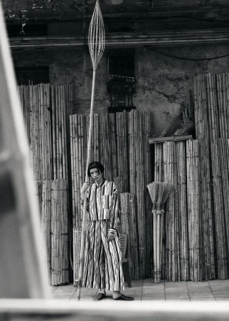 Monochrome portrait of an adult female in a striped suit holding a tall bamboo pole inside a room with bamboo walls.