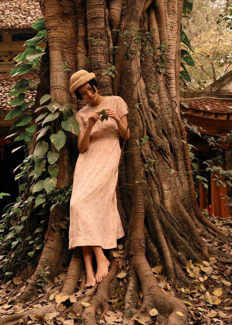 Asian woman in a dress and straw hat stands near a banyan tree, holding a leaf. Outdoor shot with natural lighting and neutral colors.