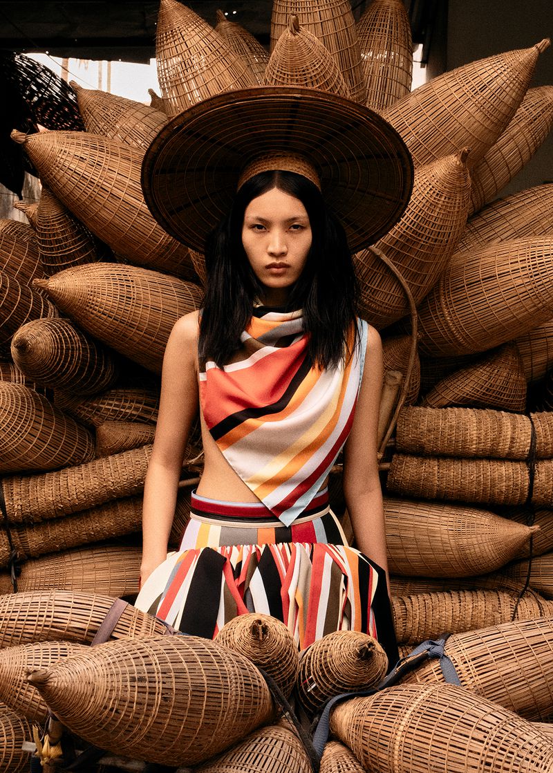 Fashion shot of a woman in a striped outfit posing in front of a stack of woven fishing traps, with a wide-brimmed hat and scarf.