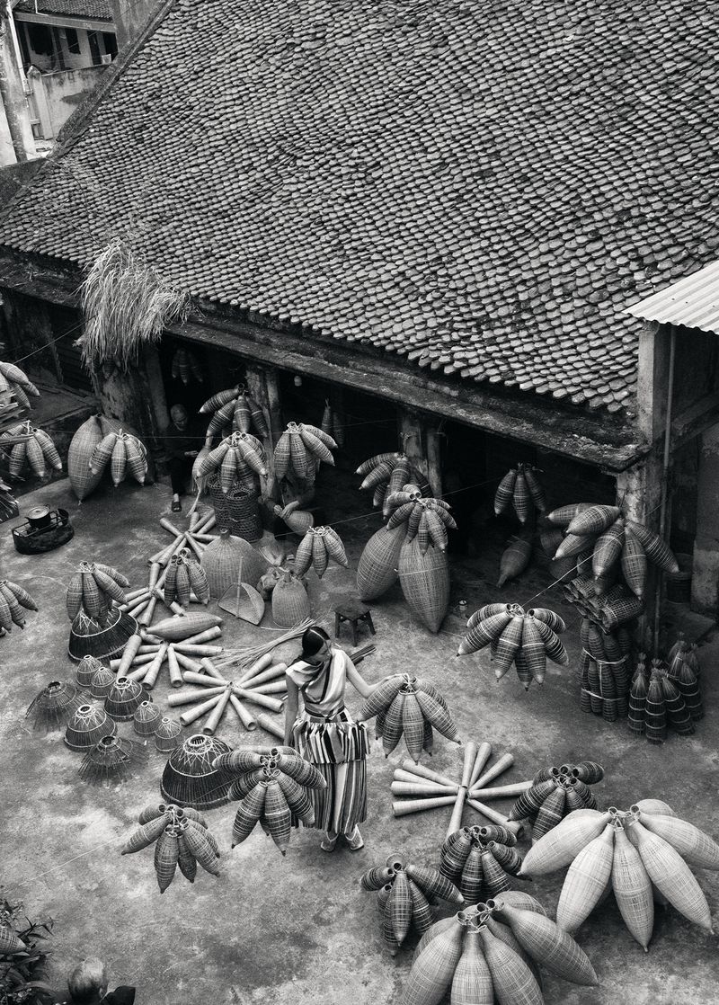 High angle shot of a woman standing in an outdoor courtyard displaying woven goods, with a tiled roof building in the background.