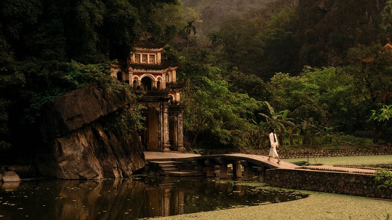 A woman in white walks across a stone bridge to an ornate building surrounded by lush greenery and a reflective pond.