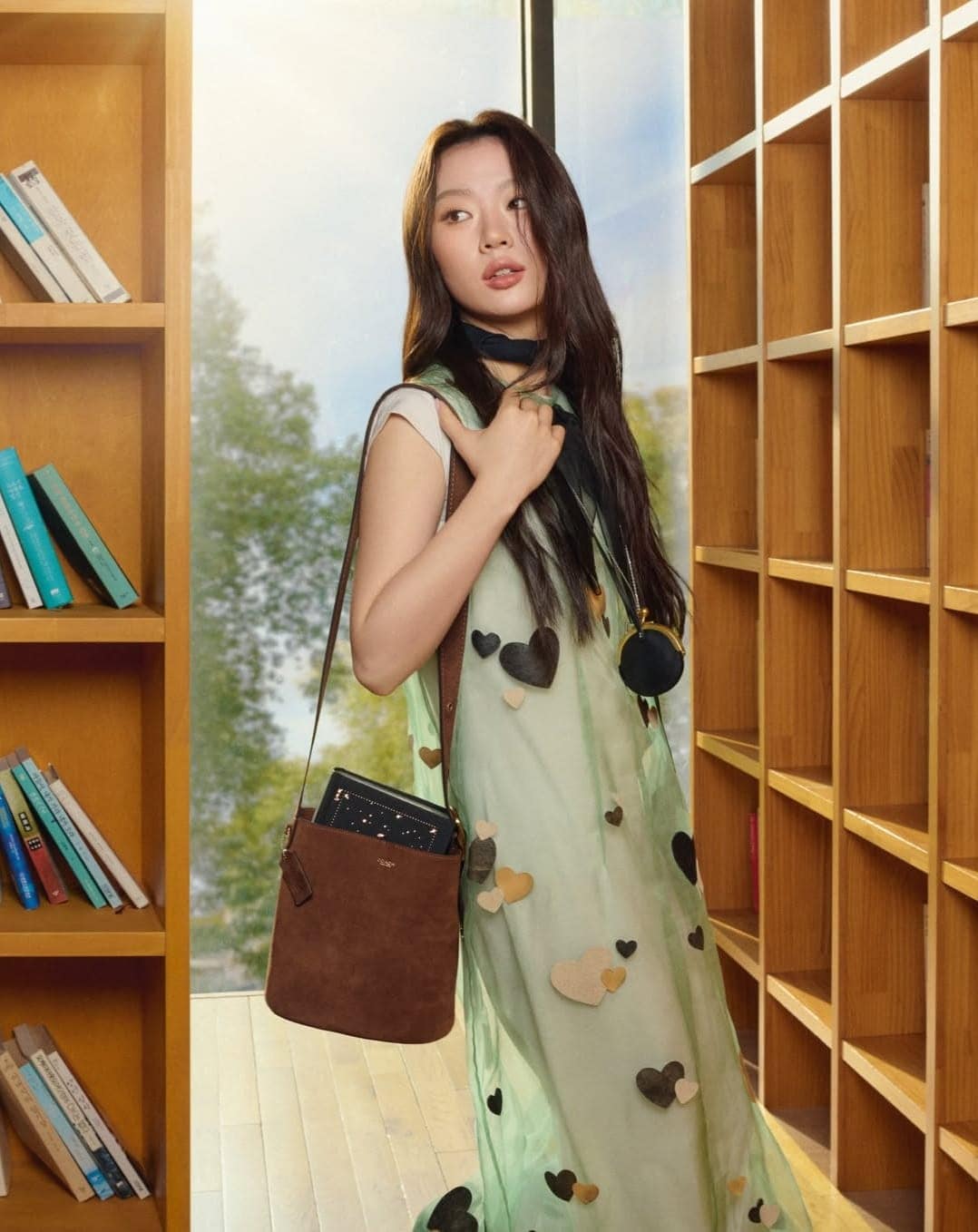 Woman wearing a sheer green dress adorned with heart appliqués and carrying a brown leather shoulder bag inside a library.