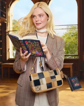 Elle Fanning holding a book and wearing a plaid blazer and patchworked leather shoulder bag inside a library