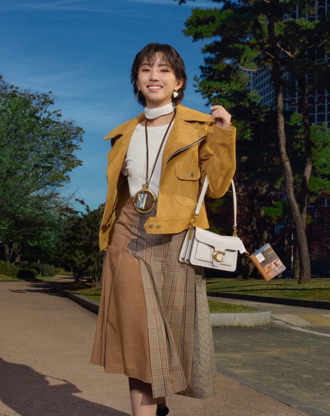 Woman in mustard yellow jacket and plaid skirt walking outdoors holding a white shoulder bag