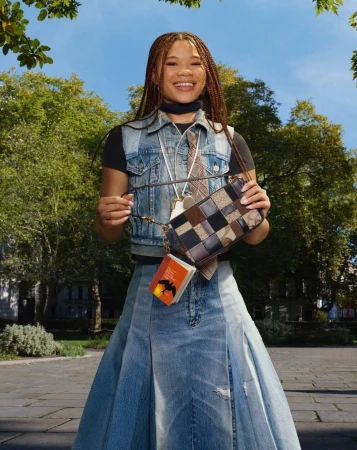 Young woman in denim vest and skirt holding patchwork handbag outdoors in park under trees