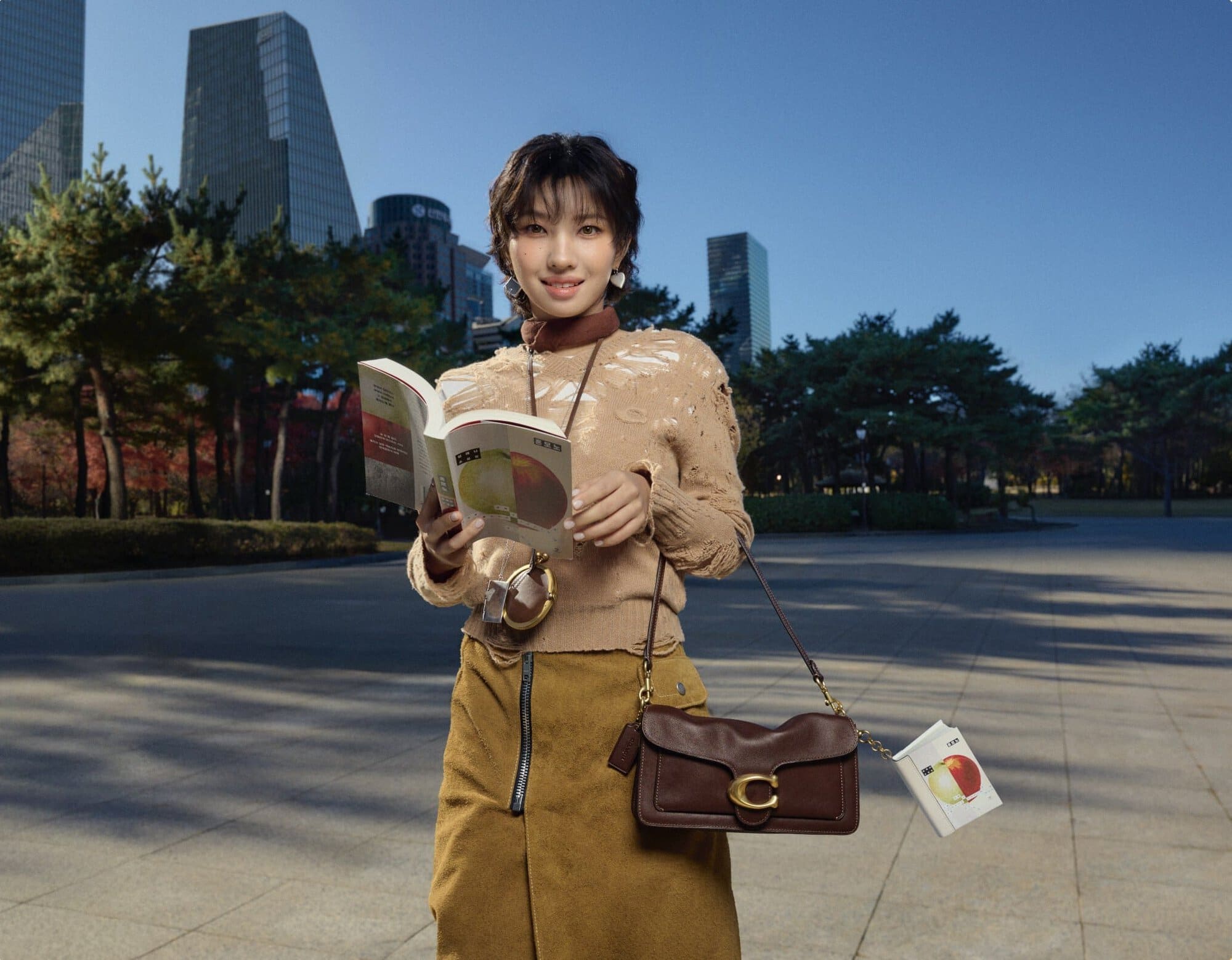 Woman holding an open book and brown leather shoulder bag outdoors in a public square with modern skyscrapers.