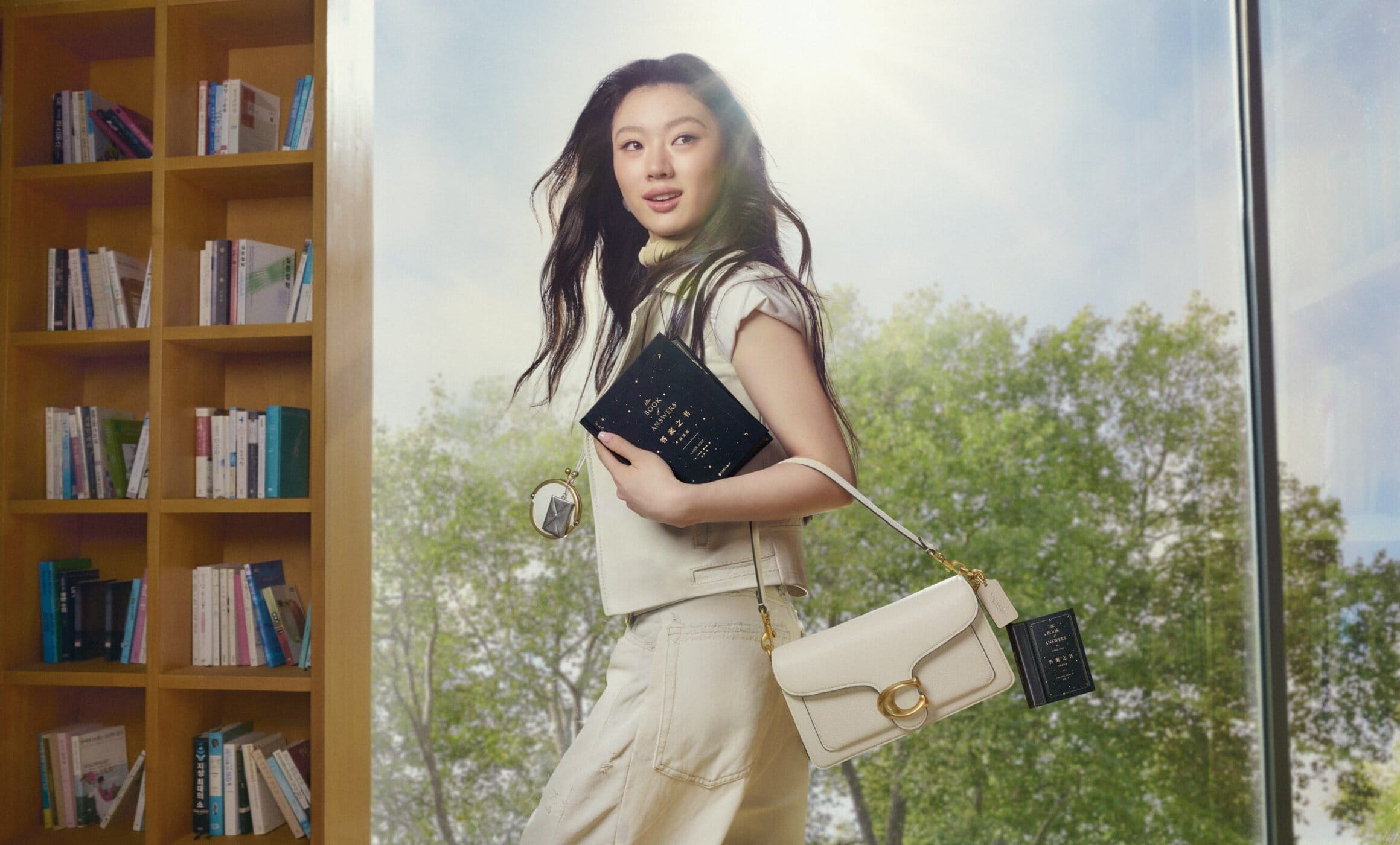 Woman holding a book and white handbag standing indoors near a large window and wooden bookshelf