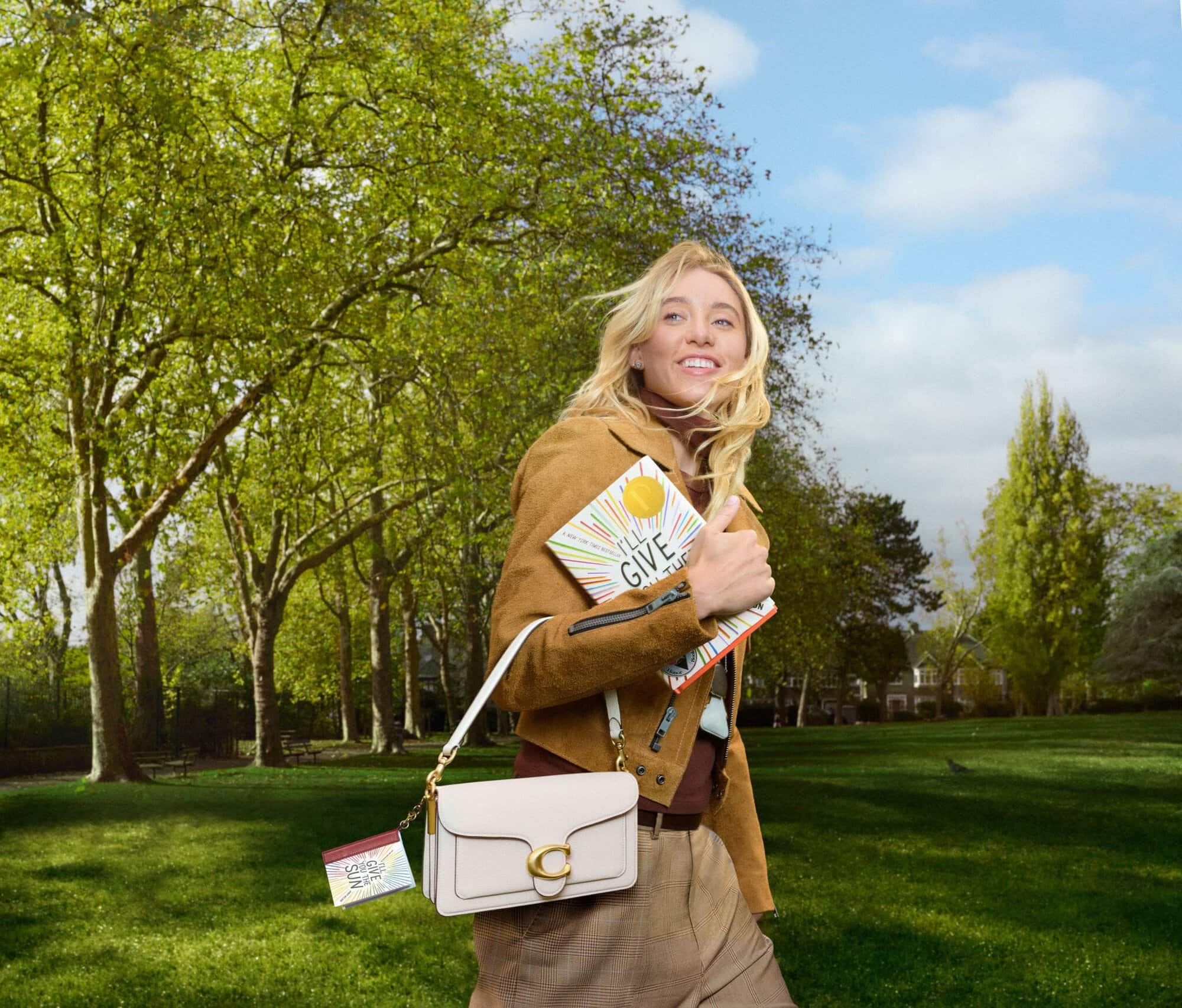 Blonde woman wearing brown jacket and carrying a white handbag walking in a park with green trees and blue sky.