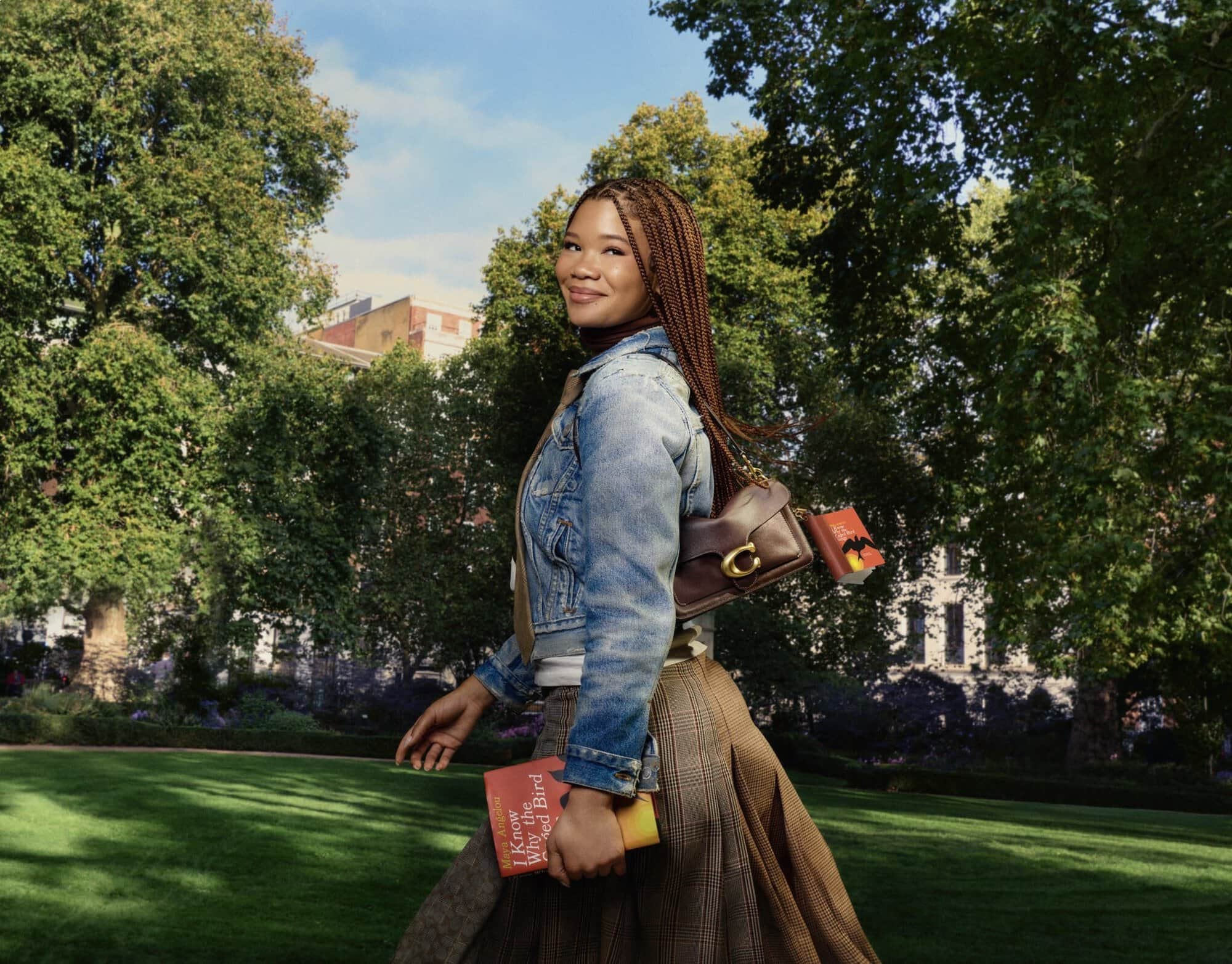 Storm Reid wearing a denim jacket and pleated plaid skirt holding a brown leather shoulder bag outdoors in a park.