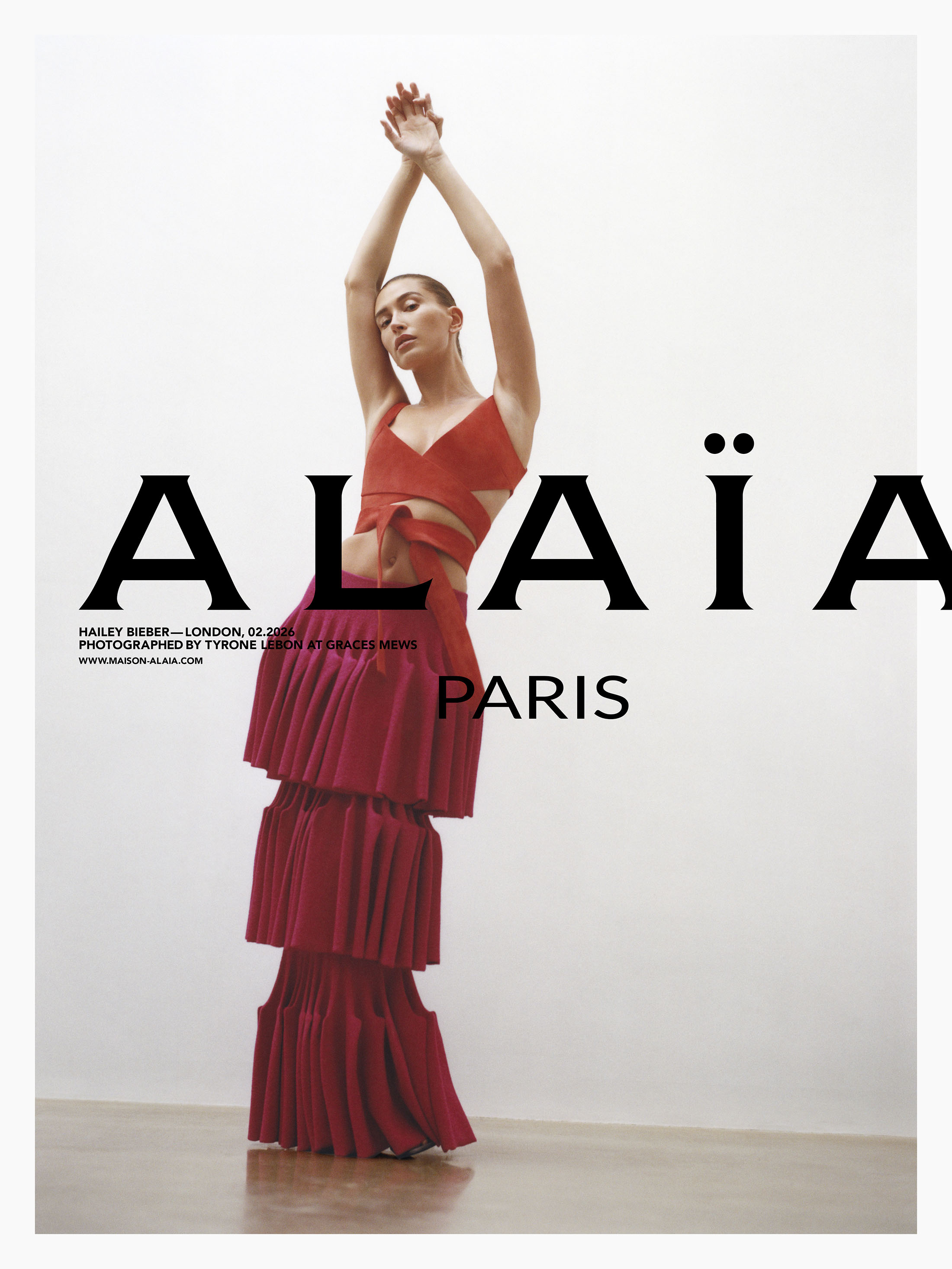 Model Hailey Bieber posing with arms raised in a red wrap top and tiered skirt against a seamless white studio background.