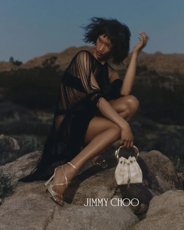 Model crouching on rock in black fringed dress holding white handbag and wearing metallic heels