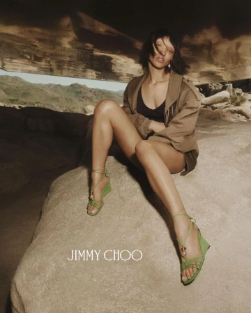 Model seated on rock wearing green strappy wedge sandals and a tan jacket for Jimmy Choo