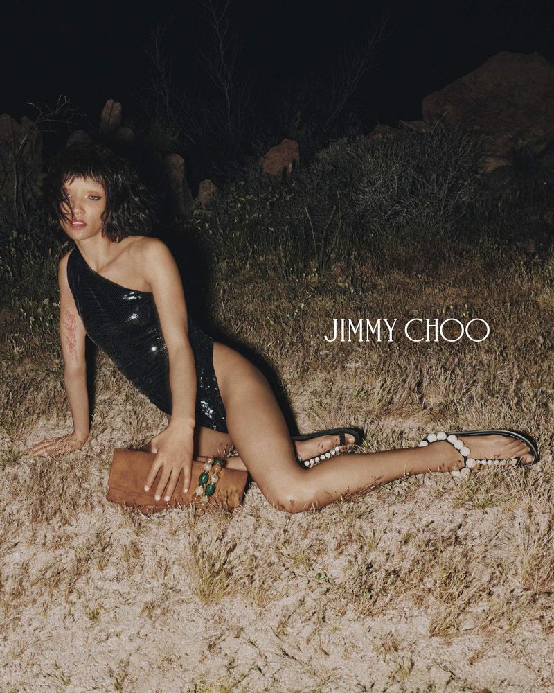 Model in a black sequined one-shoulder bodysuit and beaded sandals lying on dry grass at night for Jimmy Choo.