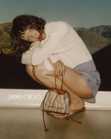 Model with dark hair crouching while holding a patterned bucket bag against a desert backdrop for Jimmy Choo Summer 2026.