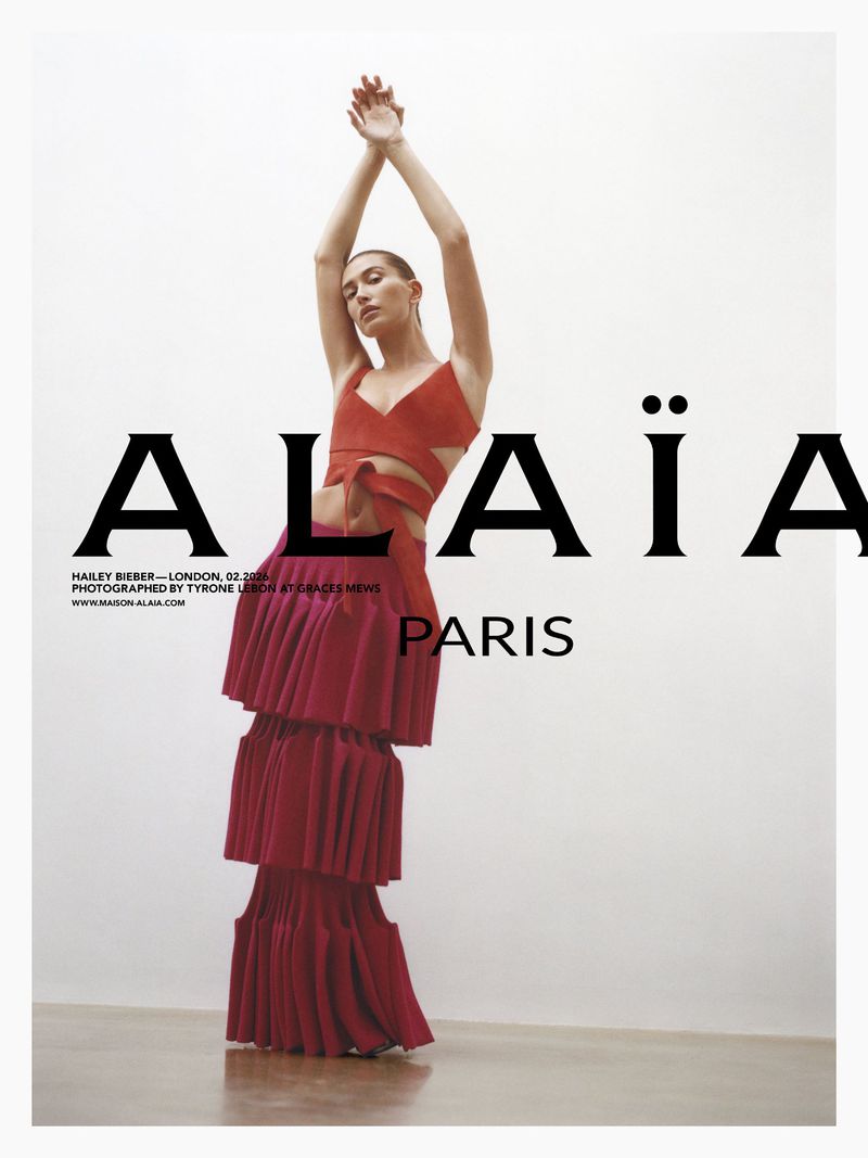 Model Hailey Bieber posing with arms raised in a red crop top and tiered pleated skirt against a white background for Alaïa