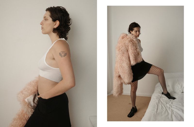 Woman in white top and black skirt poses indoors with a pink coat. Side view shows tattoo on arm. Fashion and style photography.