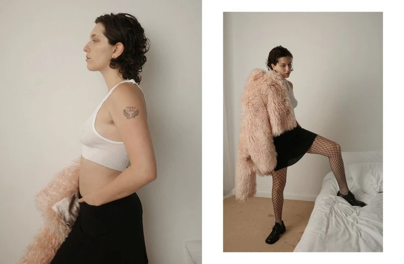 Woman in white top and black skirt poses indoors with a pink coat. Side view shows tattoo on arm. Fashion and style photography.