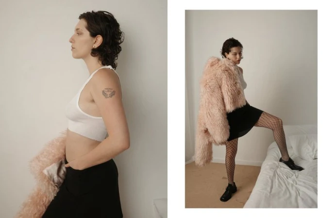 Woman in white top and black skirt poses indoors with a pink coat. Side view shows tattoo on arm. Fashion and style photography.