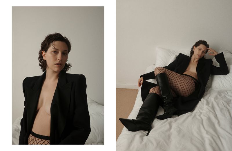 Indoor fashion shot of a young woman in a black blazer and fishnet tights lying on a bed with white sheets in soft lighting.