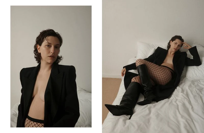 Indoor fashion shot of a young woman in a black blazer and fishnet tights lying on a bed with white sheets in soft lighting.