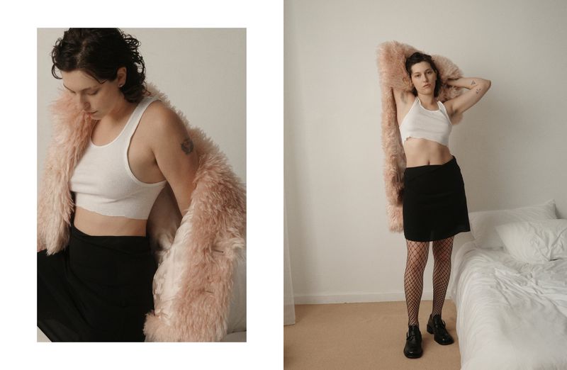 An adult woman stands in a white room wearing a crop top, skirt, and jacket, exuding a casual, fashionable, and confident vibe in a studio setting.