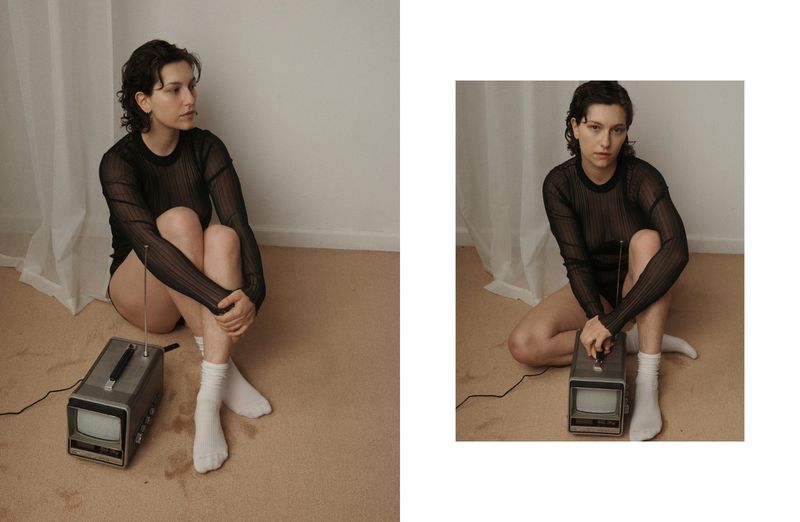 Woman with short dark hair sitting on the floor near an old TV, wearing a black top and white socks in an indoor studio setting.