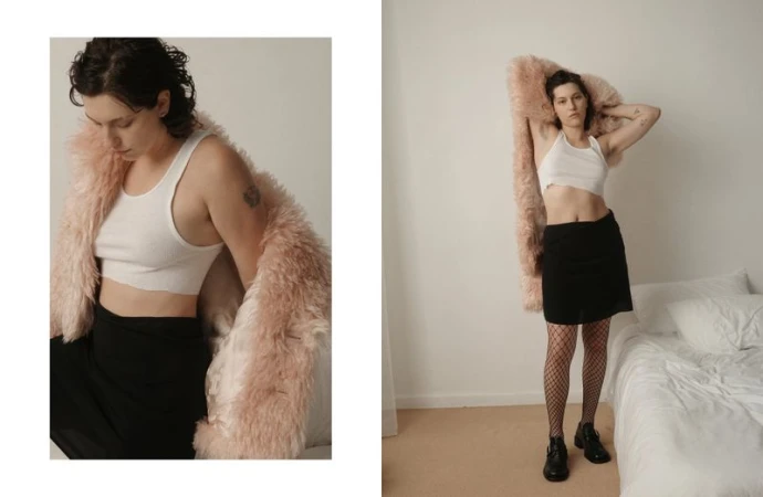King Princess posing in a cropped white tank top, black skirt, fishnets, and a shaggy pink coat beside an unmade bed.