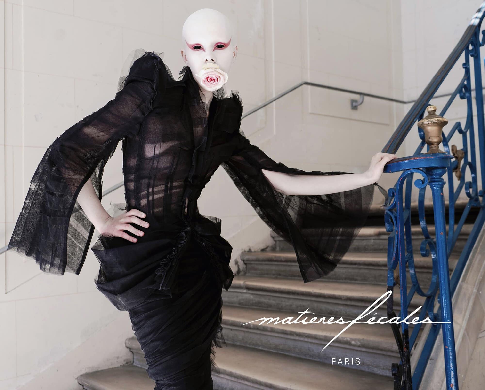Model in avant-garde black sheer ruffled dress holding a pink rose in mouth, standing on staircase with blue railing for Matières Fécales