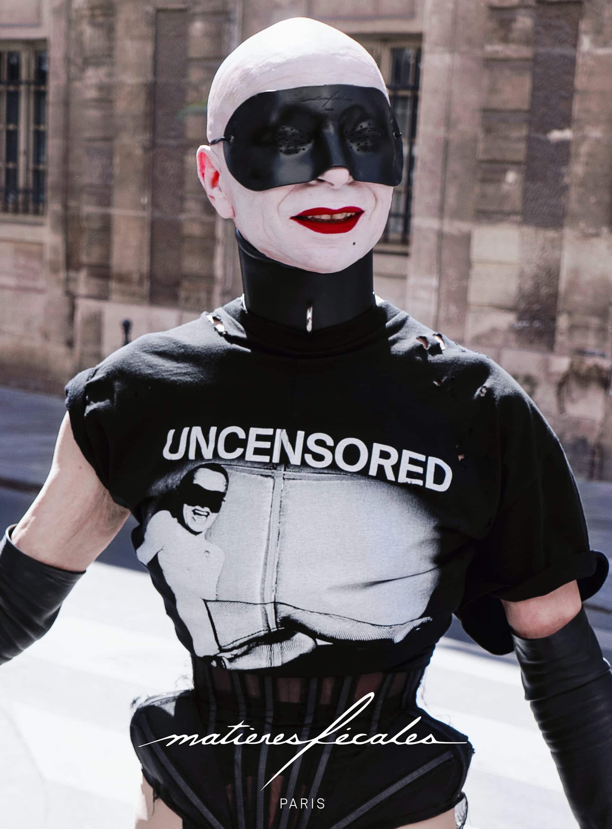 Model wearing a black uncensored graphic t-shirt, leather gloves, and dark mask standing outdoors for Matières Fécales campaign.