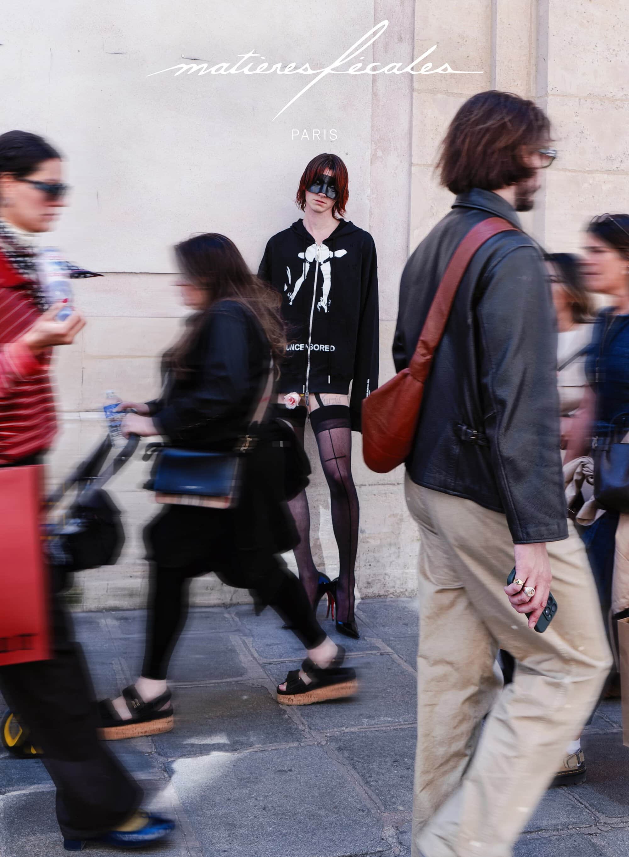 Model standing still against a stone wall while pedestrians walk past in motion blur for a Matières Fécales campaign