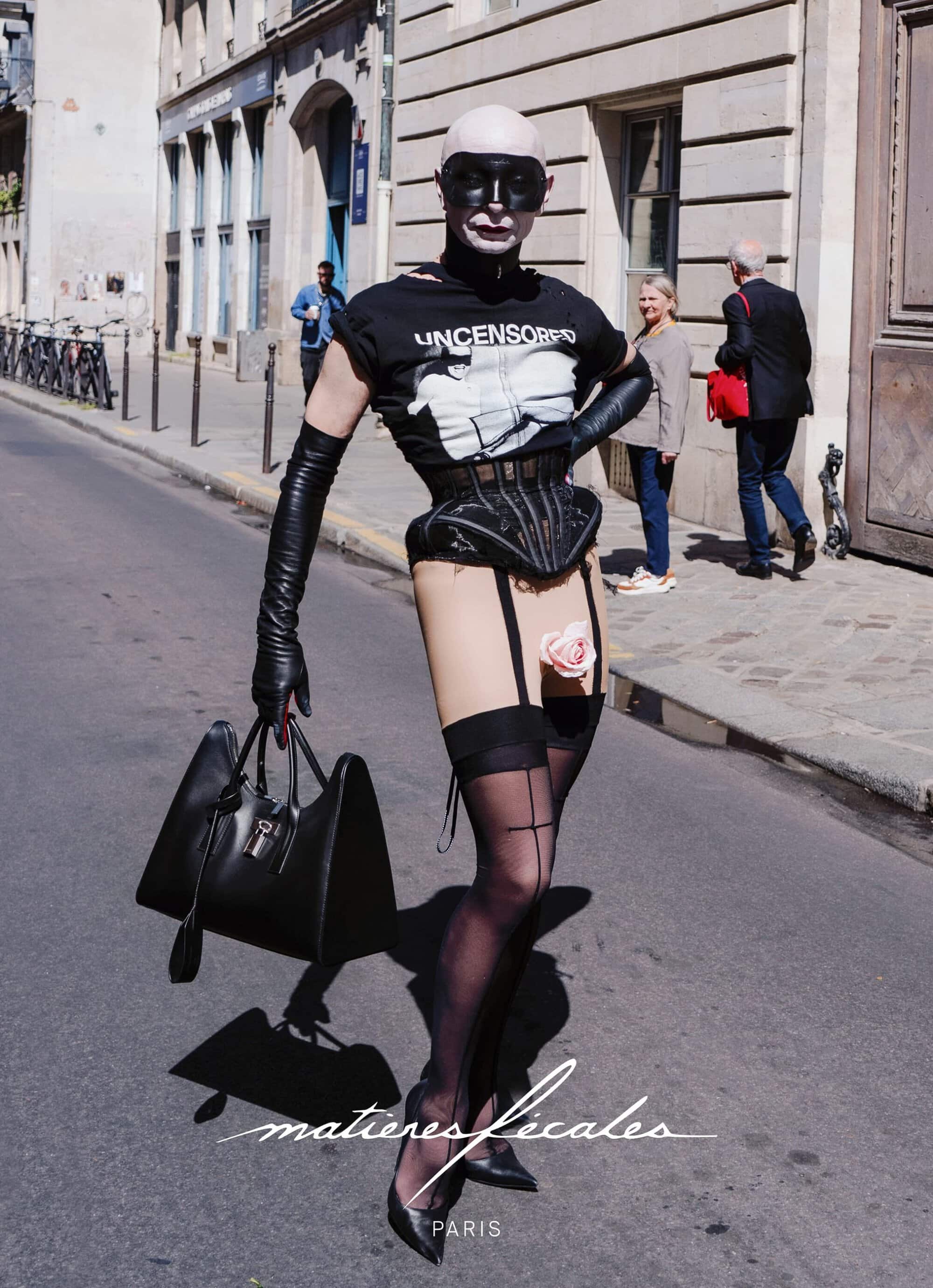 Model wearing a black corset, graphic t-shirt, and stockings holding a handbag on a sunny city street for Matieres Fecales campaign.