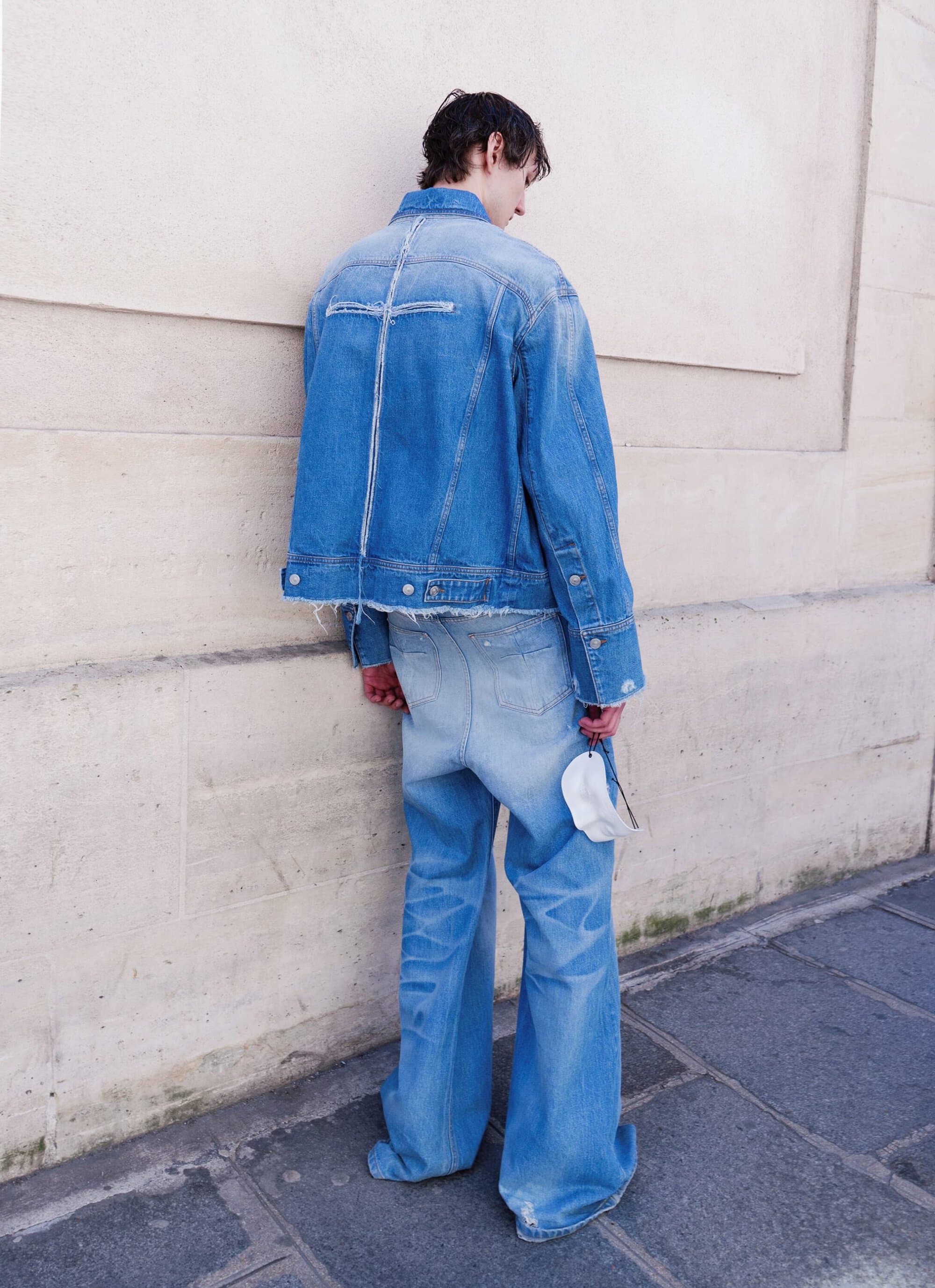 Model seen from behind wearing an oversized blue denim jacket and matching wide-leg jeans while leaning against a stone wall.