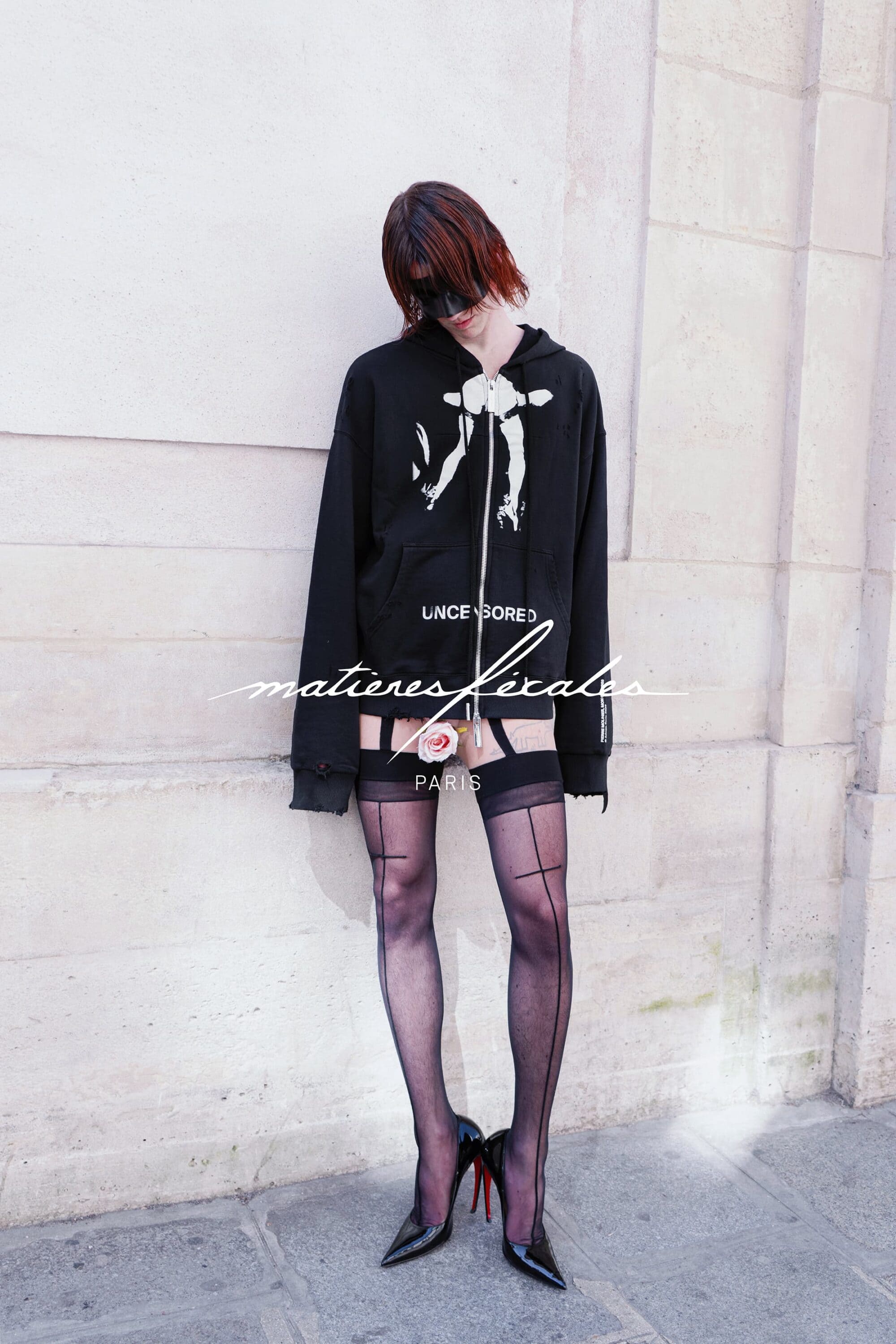 Model in black hoodie and sheer stockings wearing high heels standing against a stone wall