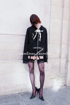 Model in black hoodie and sheer stockings wearing high heels standing against a stone wall