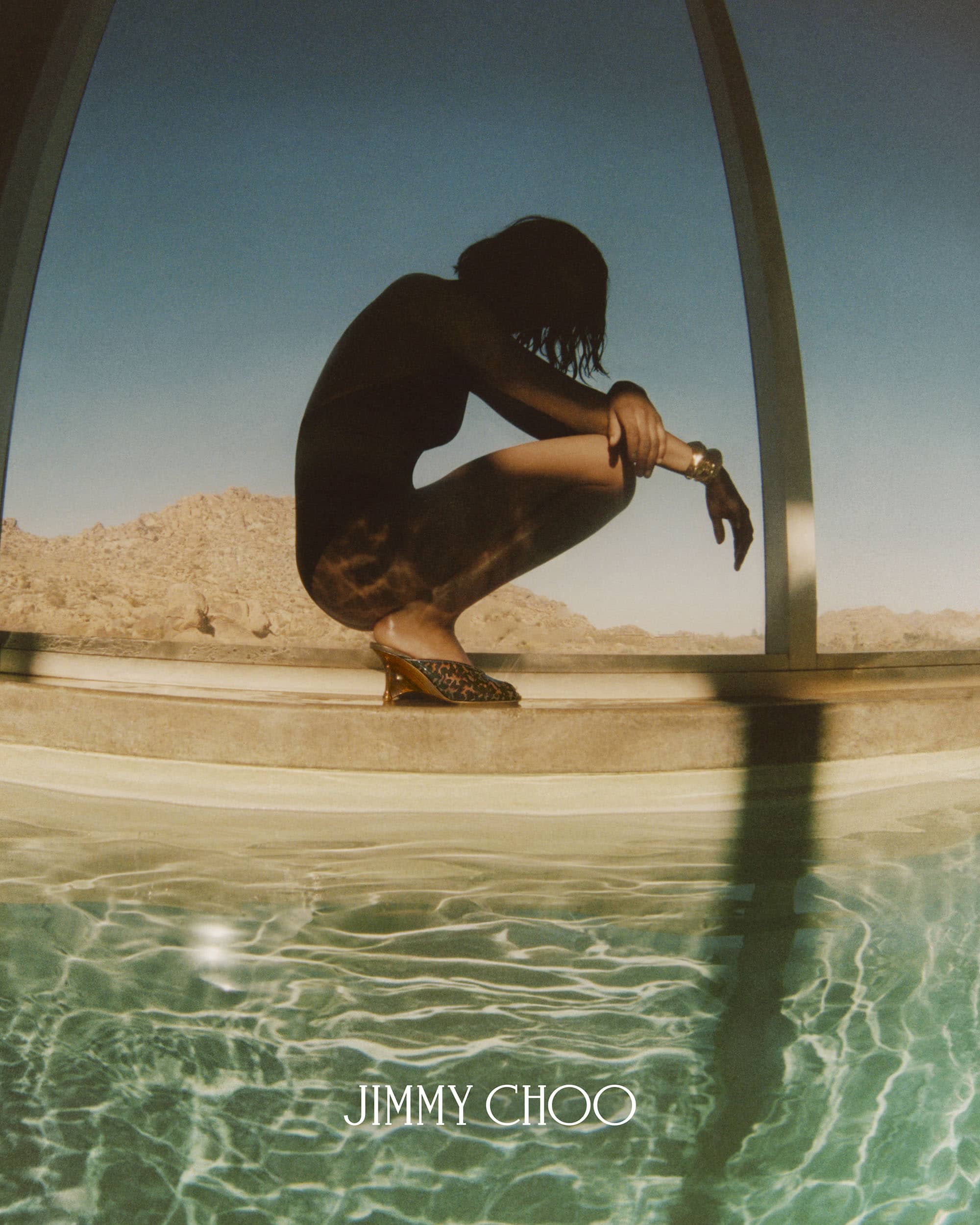Model crouching by a pool with desert and mountains visible in the background for Jimmy Choo summer campaign.