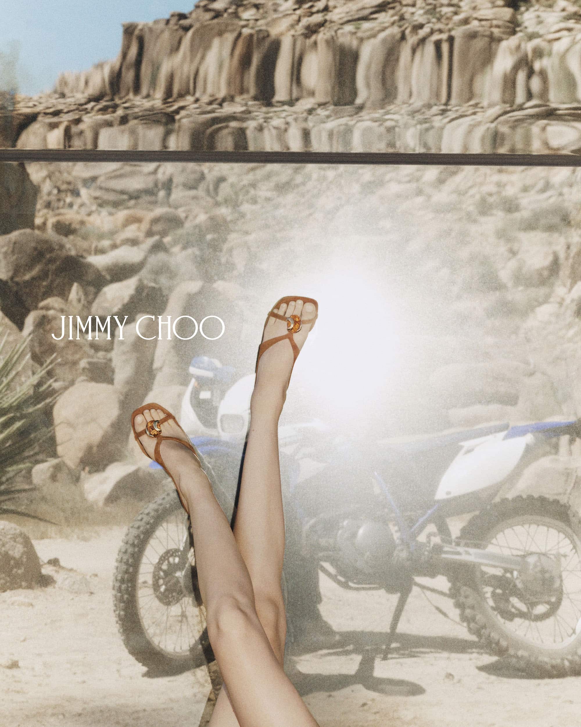 Close-up of legs wearing brown strappy sandals with a motorcycle and desert rock backdrop for Jimmy Choo.