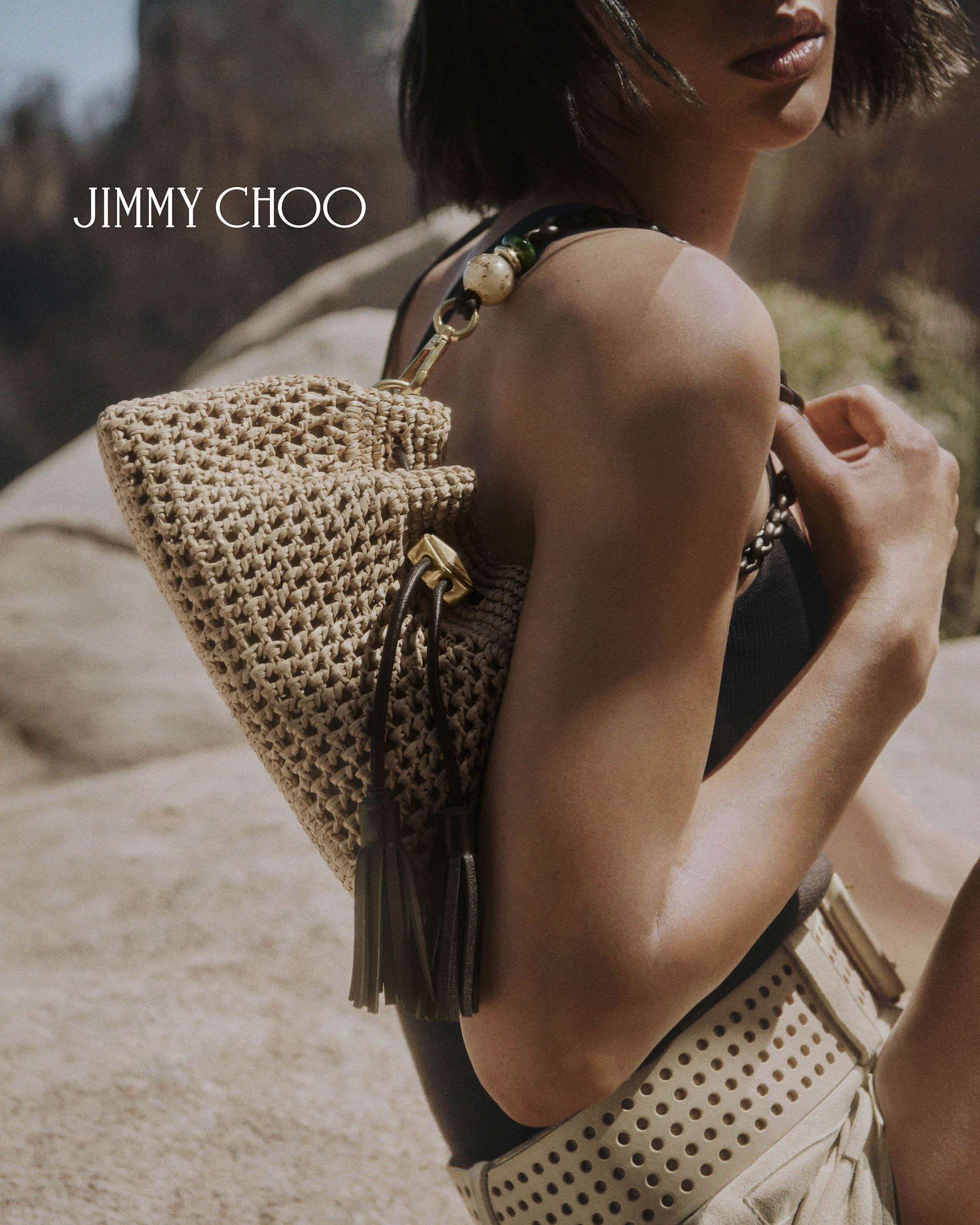Model holding a woven straw shoulder bag outdoors wearing a dark top and perforated beige shorts for Jimmy Choo.