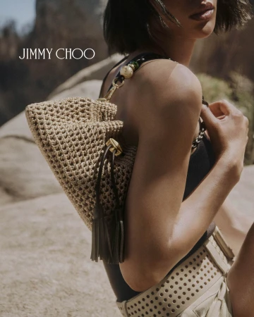 Model holding a woven straw shoulder bag outdoors wearing a dark top and perforated beige shorts for Jimmy Choo.