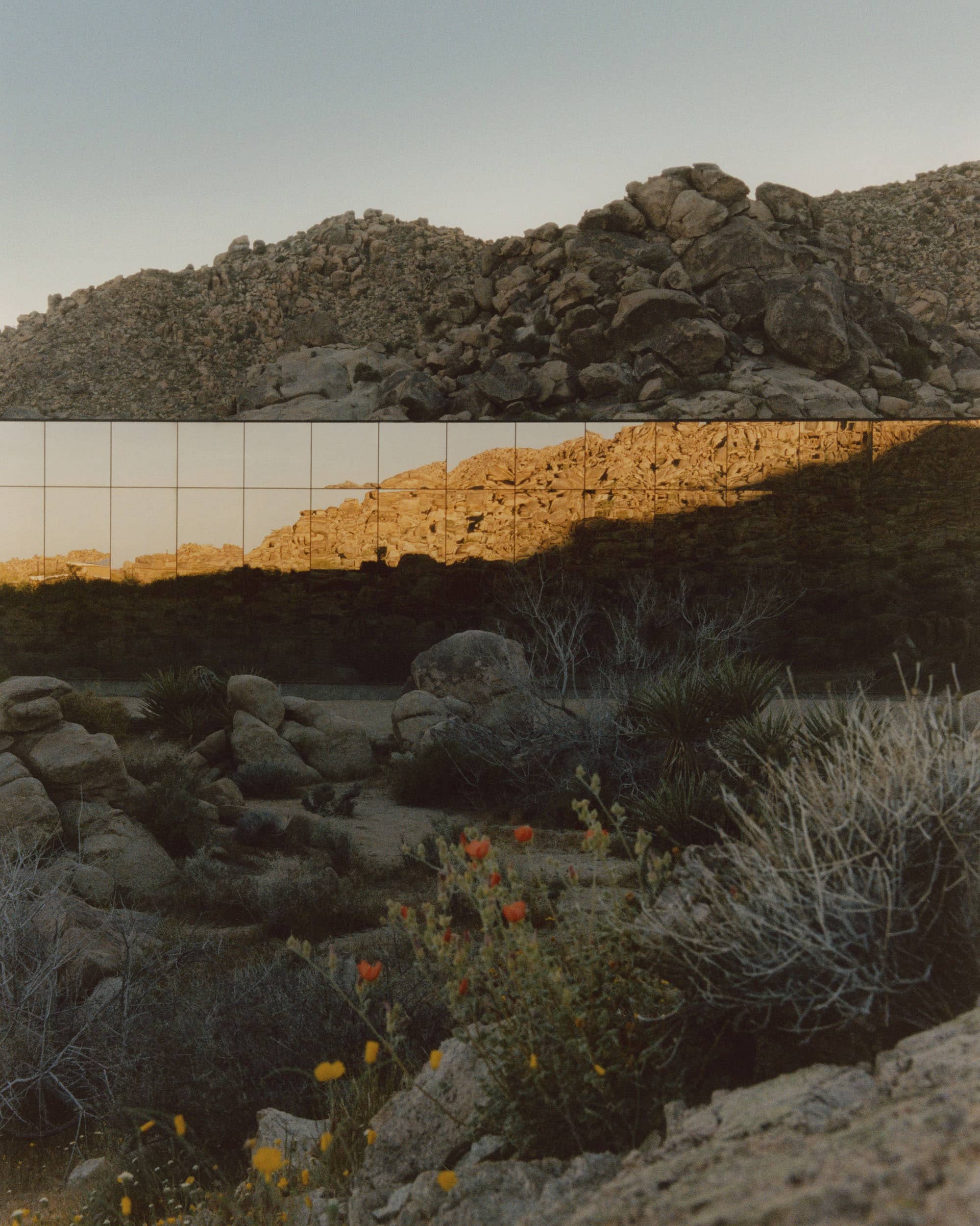 Desert landscape with rocky terrain reflecting in a mirrored glass wall under a clear sky.