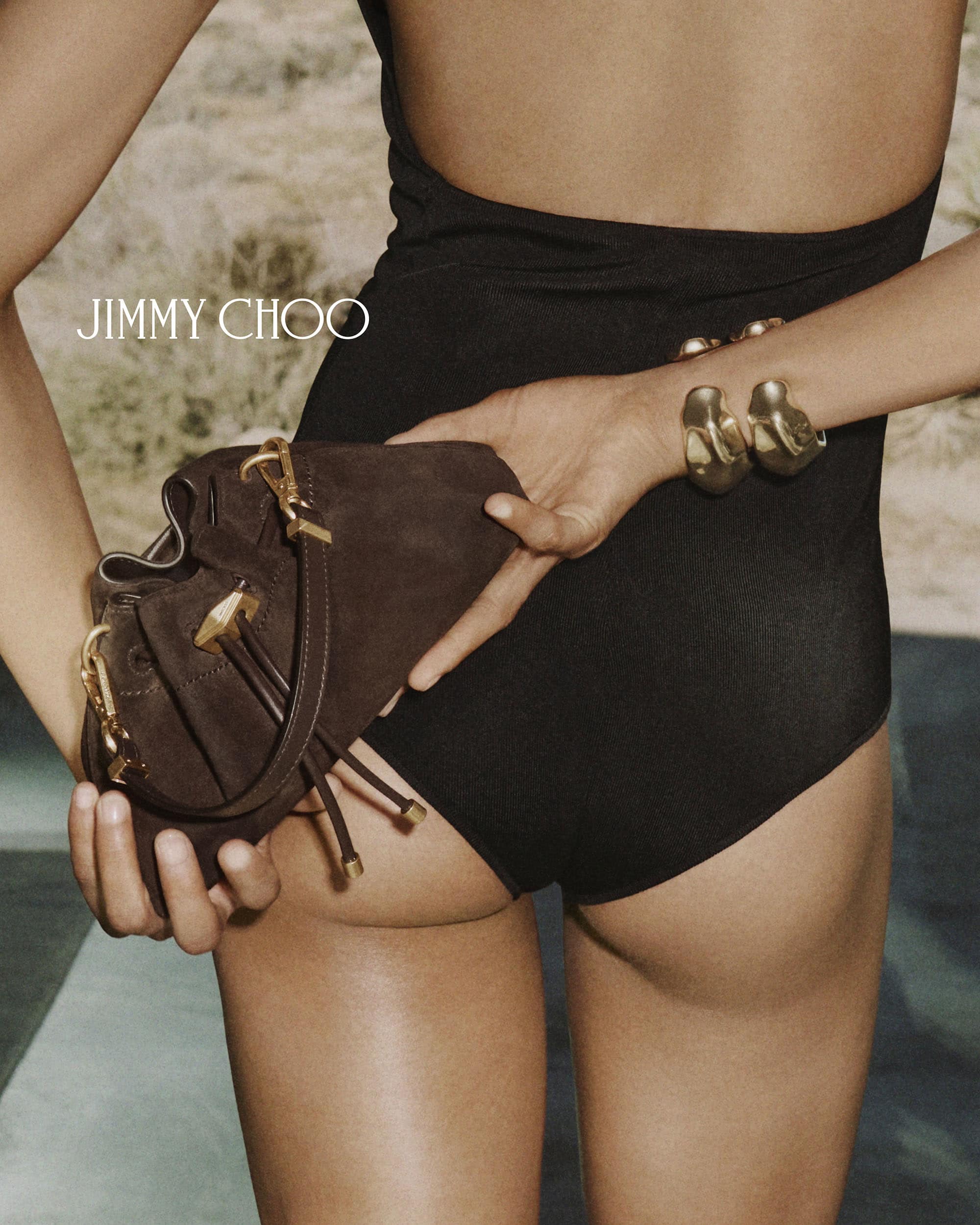 Model in black swimwear holding a brown suede handbag and wearing gold cuff bracelets outdoors